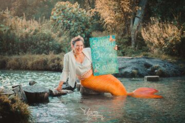 Hanna als Seejungfrau mit Blazer sitzt im Wasser. Sie lächelt in die Kamera. Im Hintergrund sind Bäume. Hanna hält ihr Schreib-deinen-Roman-Schild.