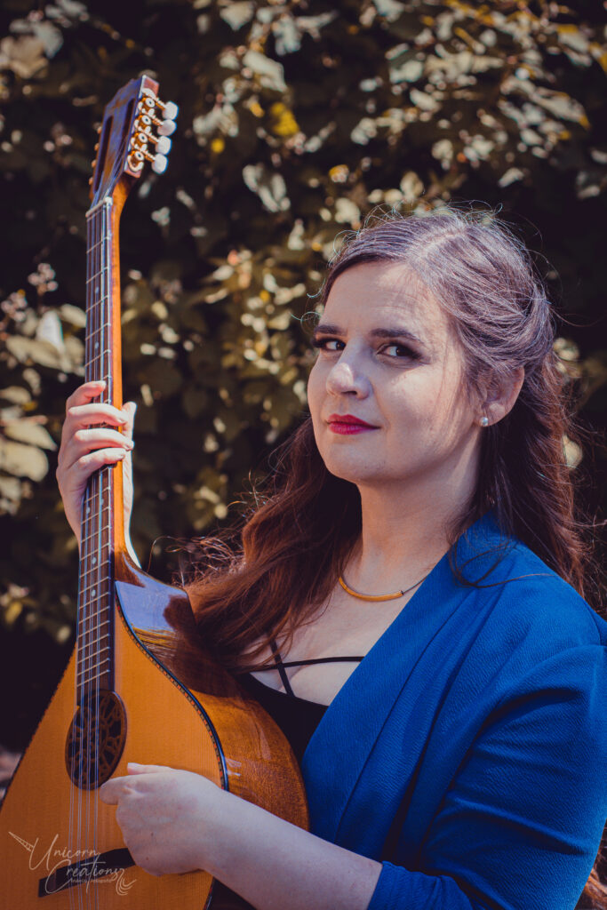 Hanna steht mit petrolfarbenem Blazer vor einer Hecke und hält eine Bouzouki, ein griechisches Saiteninstrument.